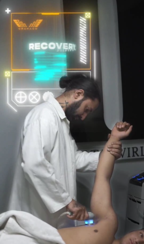 A person in a white coat performing a recovery treatment on a shirtless individual using a handheld device under their arm, with a glowing digital recovery interface in the background.