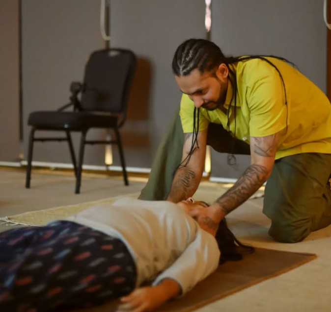 Man in yellow shirt with braided hair kneeling while adjusting a woman's neck as she lies on a yoga mat.