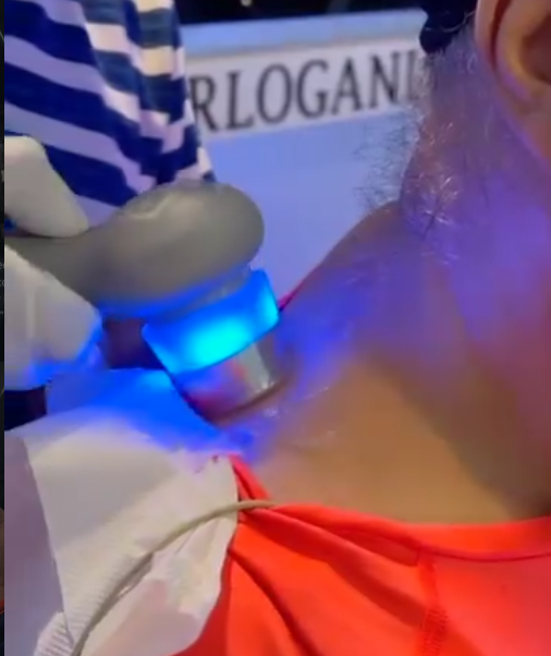 Gloved hand holding a device emitting blue light applied to a person's neck near the shoulder.