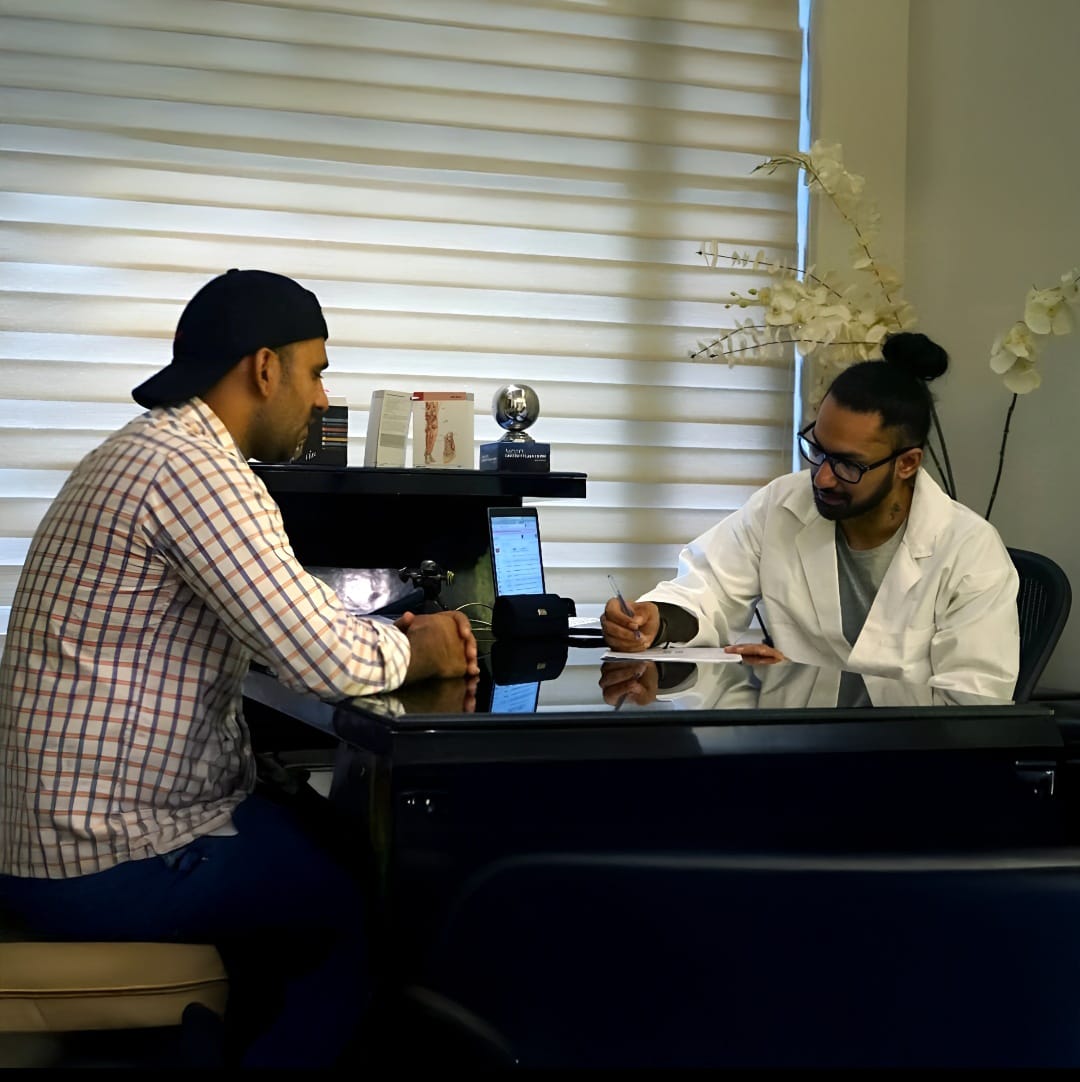 A man in a white coat is writing on a clipboard while talking to a man in a plaid shirt and black cap sitting across a desk.
