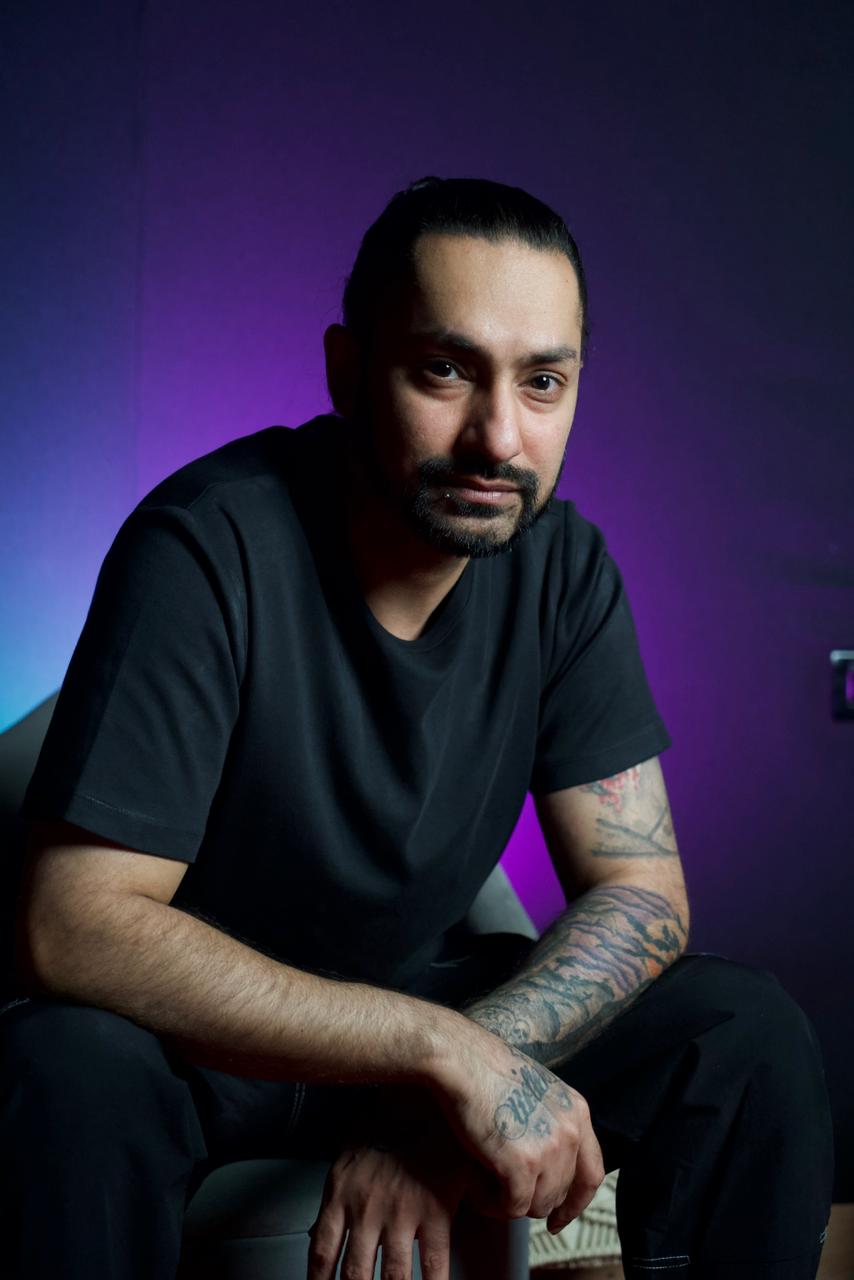 Man with a beard and tattoos sitting on a chair against a purple and blue gradient background.